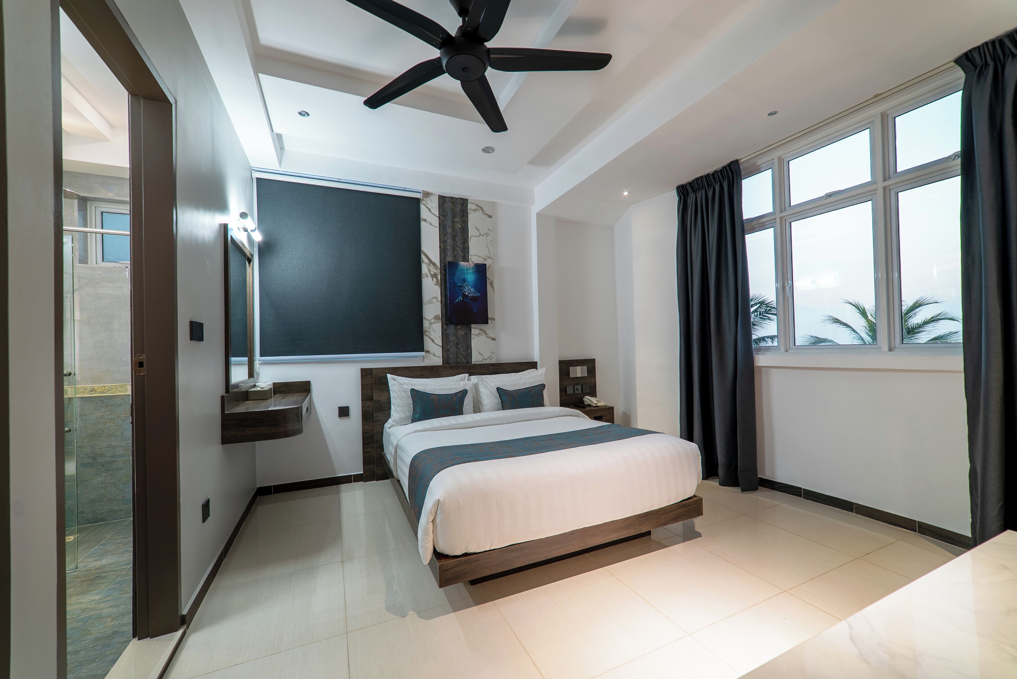Een moderne hotelkamer met een bed, een plafondventilator, een groot raam met gordijnen, een wandtelevisie en een accentmuur van marmer.