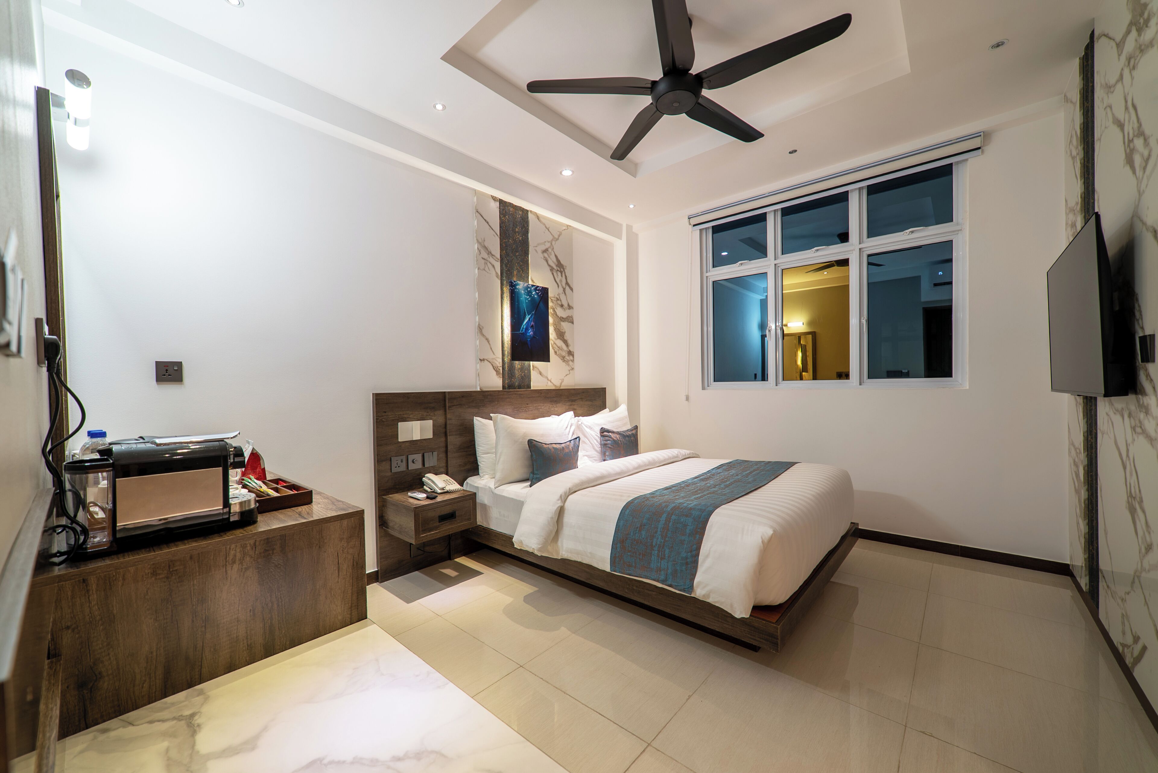 A modern hotel room with a bed, a desk, a ceiling fan, a large window, and a TV.