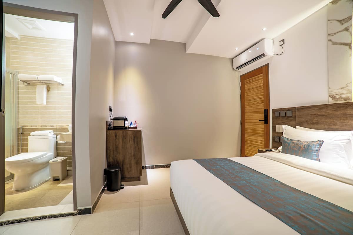 A hotel room with a bed, a wooden headboard, an air conditioning unit, a small desk with a phone, and a bathroom with a toilet and towel rack.