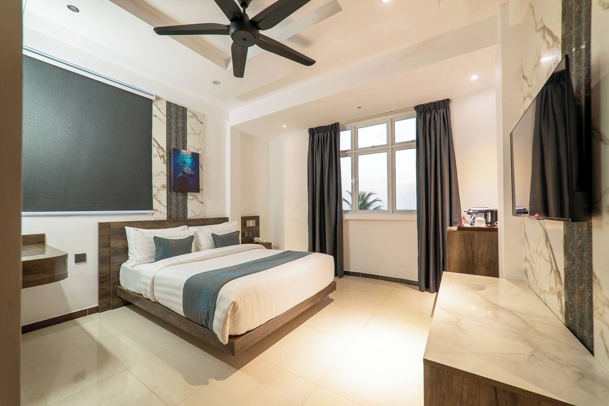 A modern hotel room with a bed, a ceiling fan, a TV, a window with curtains, and a marble countertop.