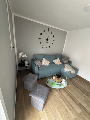 Un salon avec un canapé bleu, une table basse ronde en verre, un pouf gris, une horloge murale et une petite table d’appoint sur laquelle est posée une plante.
