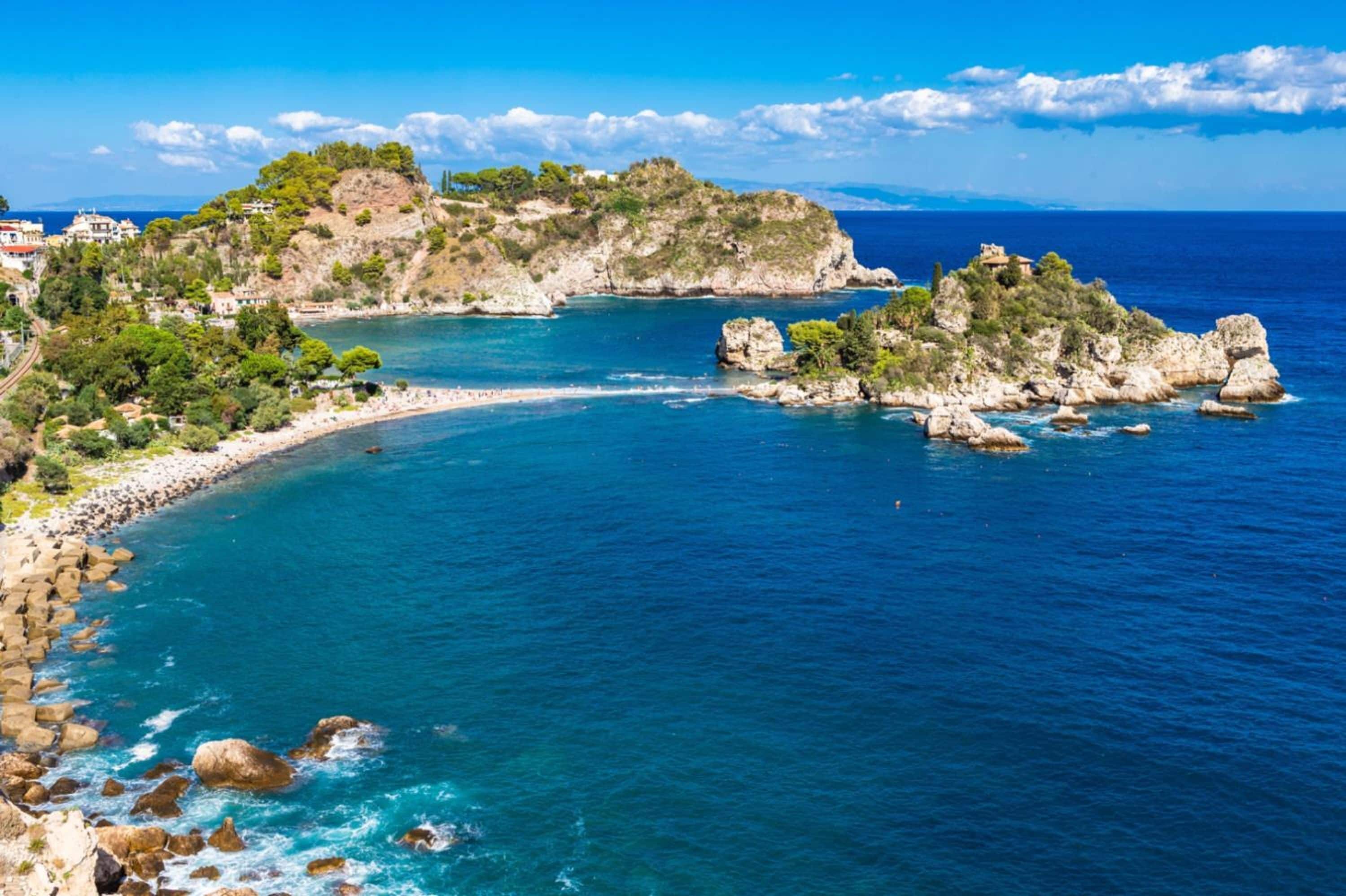 A coastal landscape with rocky terrain, clear blue waters, and buildings on the hills.