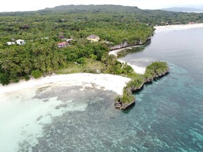 An aerial view of a coastal area with a beach, clear waters, and dense greenery.
