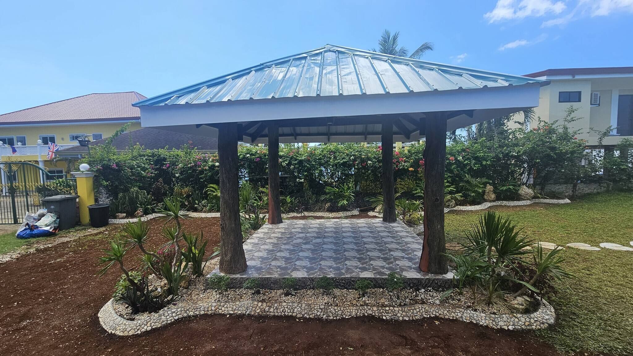 Un gazebo con pavimento in tegole e tetto in metallo, circondato da un giardino con diverse piante e un cordolo in ghiaia.
