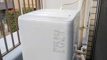 A white washing machine on a balcony with a metal railing and a building in the background.