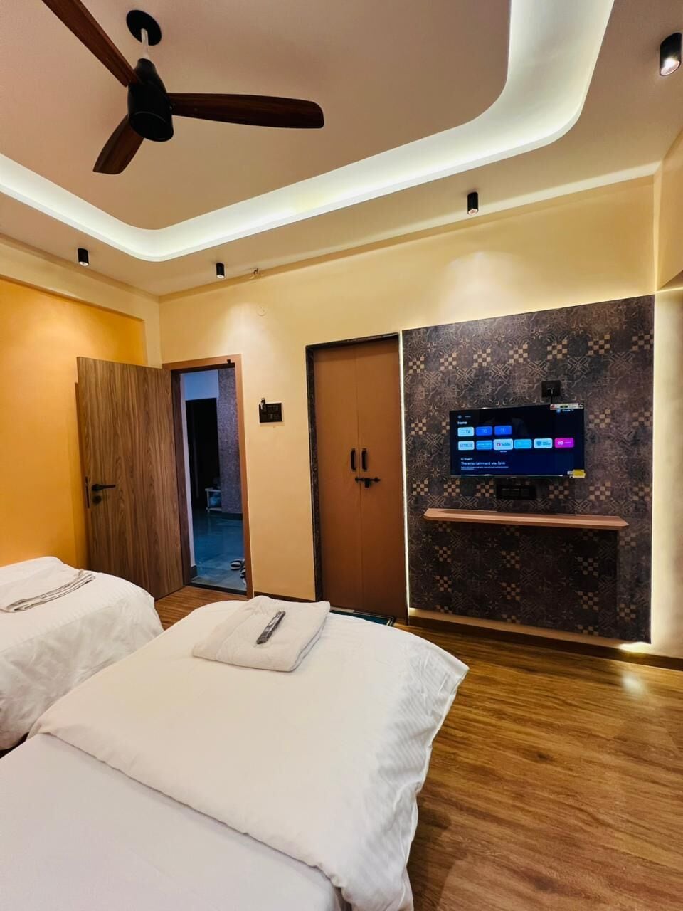 A hotel room with two beds, a ceiling fan, a TV mounted on a patterned wall, and a wooden wardrobe.