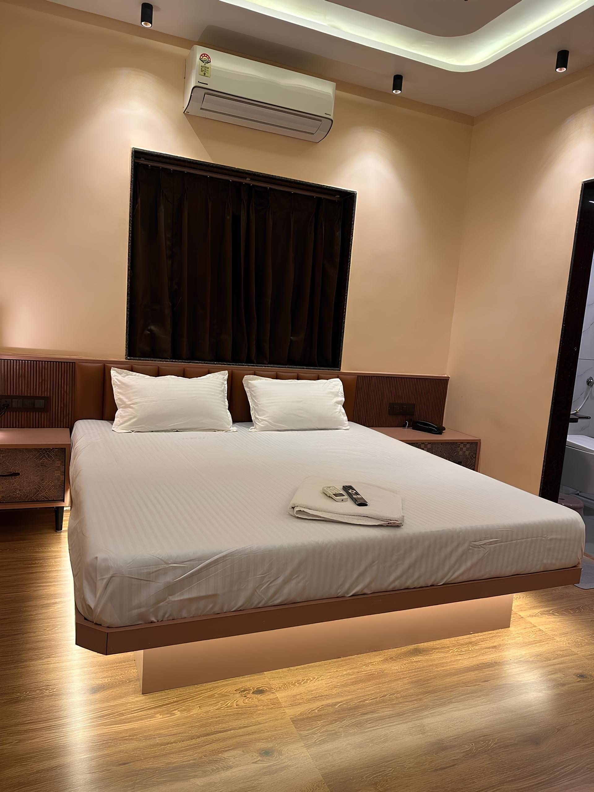 A modern hotel room with a large bed, a bedside table, an air conditioning unit, and a window with curtains.