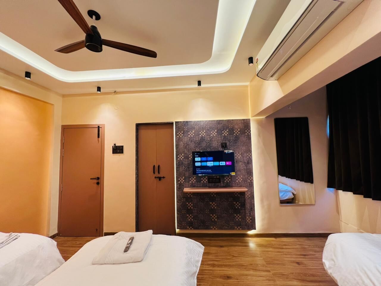 A hotel room with two beds, a ceiling fan, an air conditioning unit, a flat-screen TV mounted on the wall, and a mirror.