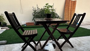 A patio with a table and two chairs, a small dog, and a garden with plants and a bamboo screen. - Olivia's BNB: Fully furnished private suite in Jimi Ameriya (Al Ain)