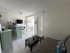 Een moderne woonkamer met een bank, een salontafel en een televisie. Er is een keukenhoek met een koffieapparaat en een eettafel met stoelen.