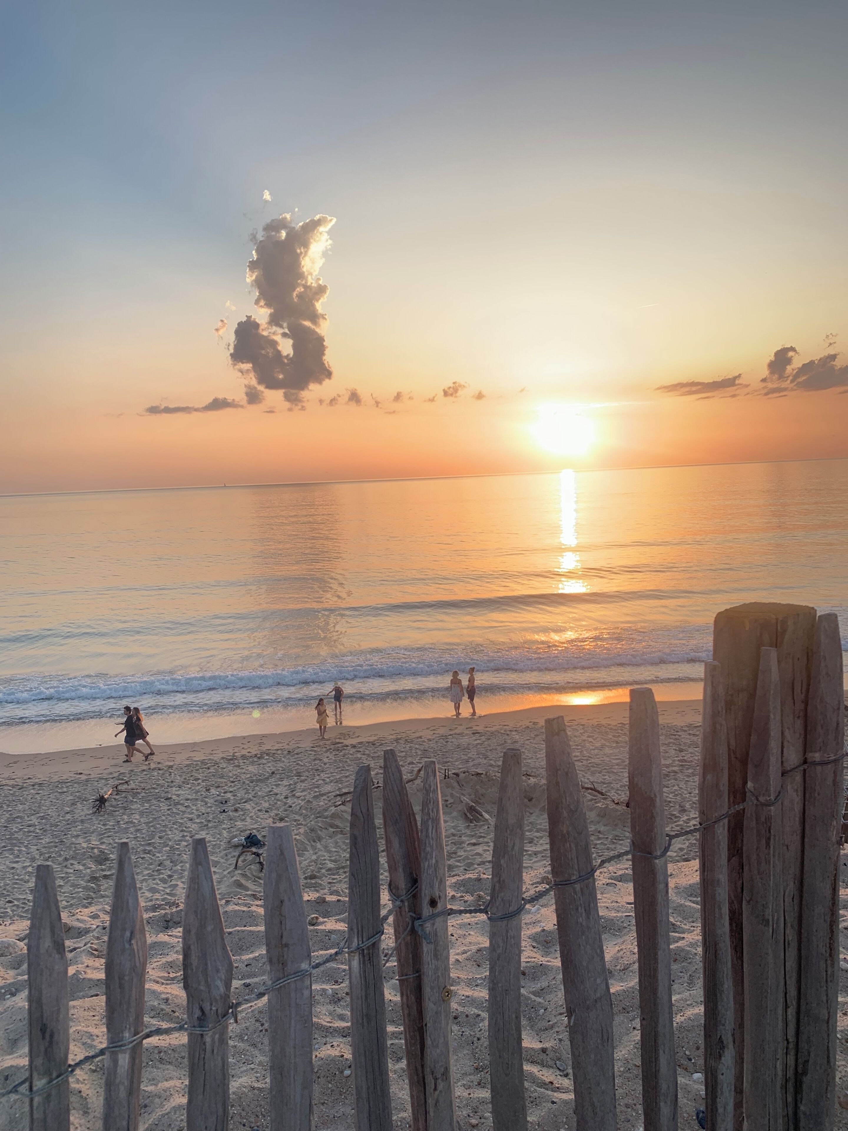 A beach scene with a wooden fence in the foreground, a sunset in the background, and people walking along the shore.