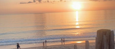 Une scène de plage avec une clôture en bois au premier plan, un coucher de soleil en arrière-plan et des personnes se promenant le long du rivage.