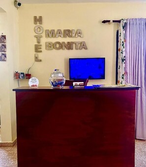 Ein Hotelrezeptionstresen mit einer Namensschild, auf der 'HOTEL MARIA BONITA' steht, einem Fernseher, einer dekorativen Vase und einem kleinen Schild an der Wand.