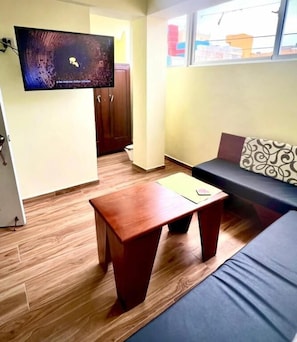 Una habitación con piso de madera, un sofá con cojines estampados, una pequeña mesa de madera, un televisor montado en la pared y una puerta que da a otra habitación.