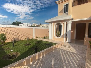A building with a beige exterior, a paved courtyard with a patterned floor, and a small garden area with a flower. - Large house with AC WiFi parking  Ideal for families (San Juan de la Maguana)