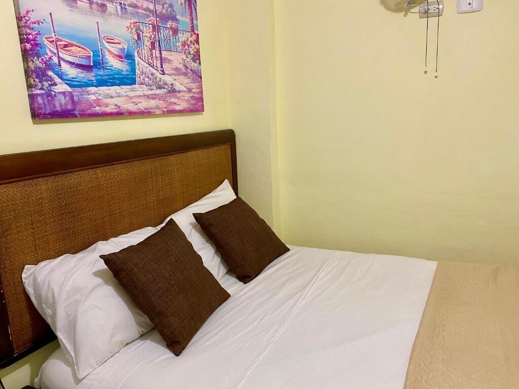 A bed with white bedding and two brown pillows, a wooden headboard, and a painting of boats on the wall.