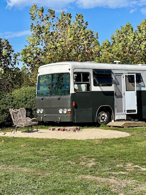 Een stilstaande bus met een deur en ramen, een rieten stoel, en een grasveld met bomen op de achtergrond.