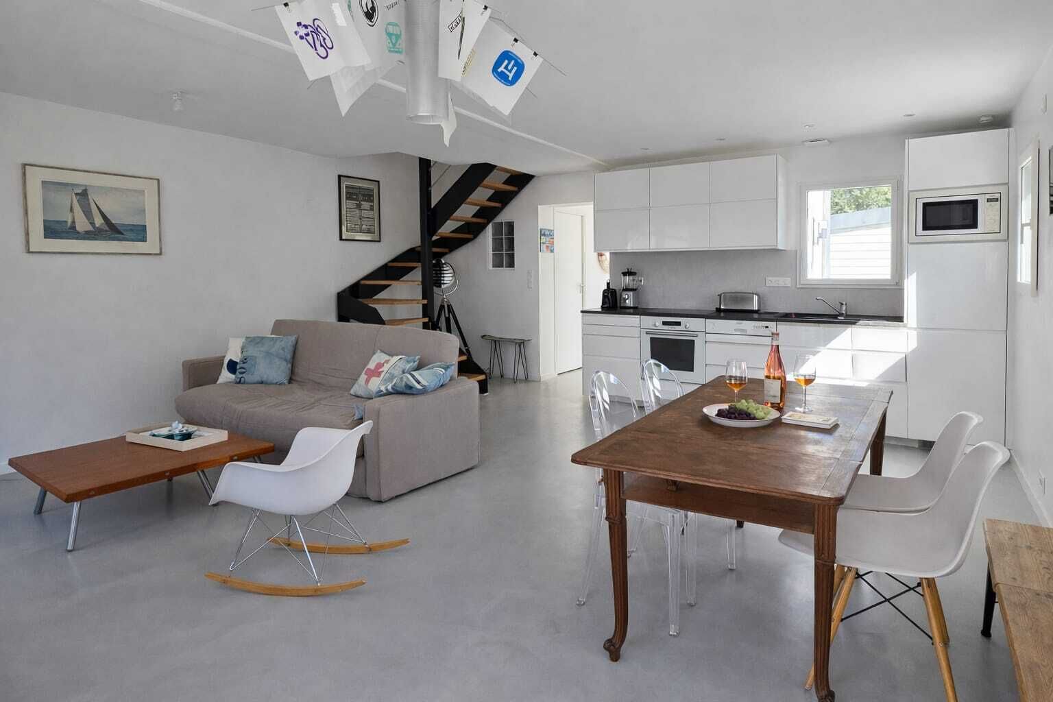 A modern living space with a sofa, dining table, chairs, a staircase, kitchen area, and a view of the outdoors.