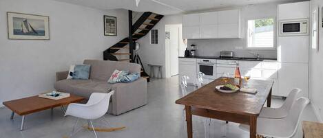 A modern living space with a sofa, dining table, chairs, a staircase, kitchen area, and a view of the outdoors.