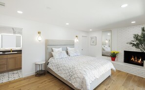 Ein modernes Schlafzimmer mit einem großen Bett, einem Nachttisch, einem Spiegel, einem Kamin und einem kleinen Beistelltisch.