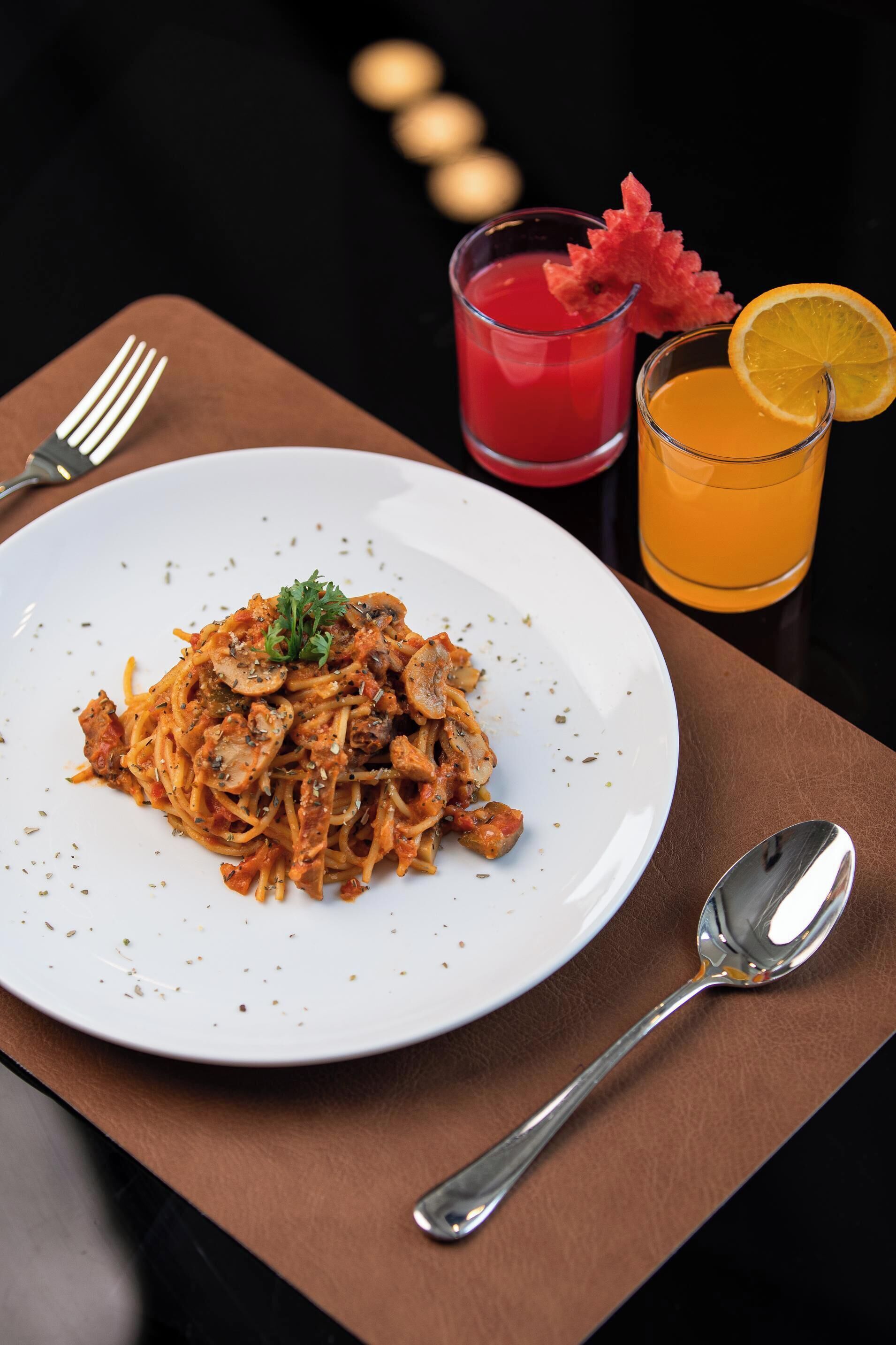 A plate of spaghetti with a garnish, a glass of red drink, and a glass of orange juice on a table.