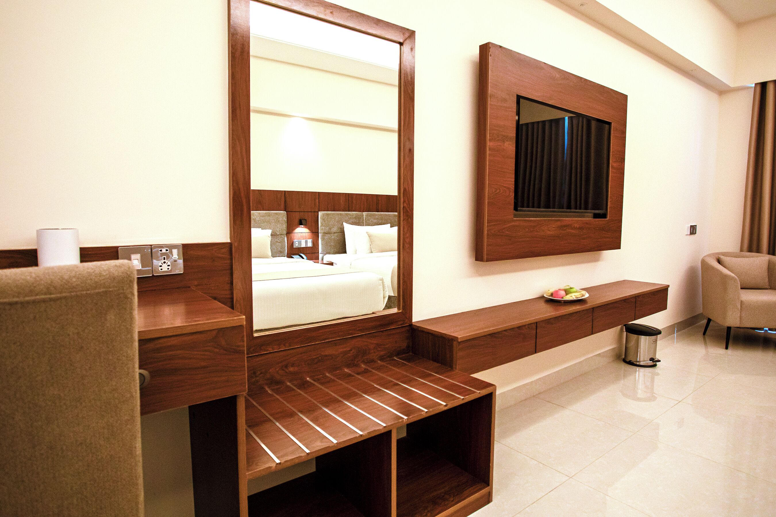 A hotel room with a large mirror, a bed with a headboard, a wooden console table, a small trash bin, and a chair.