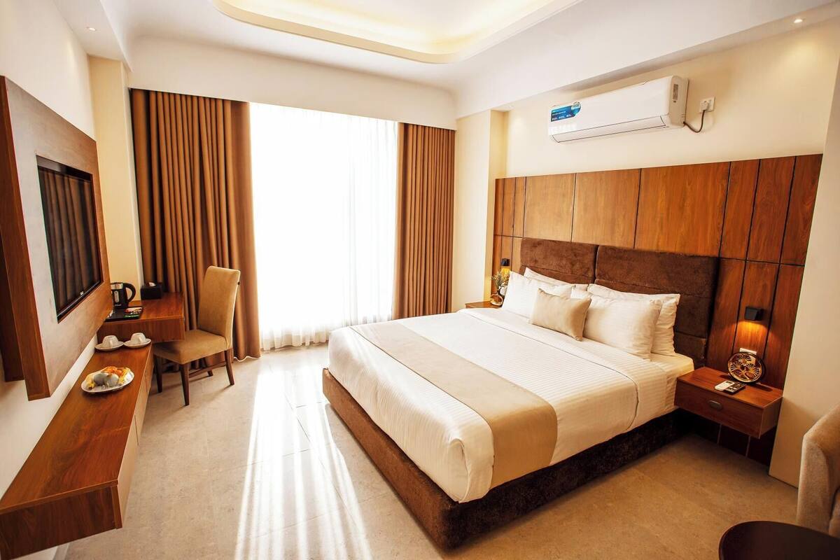 A hotel room with a large bed, a desk, a chair, a TV, and an air conditioning unit.