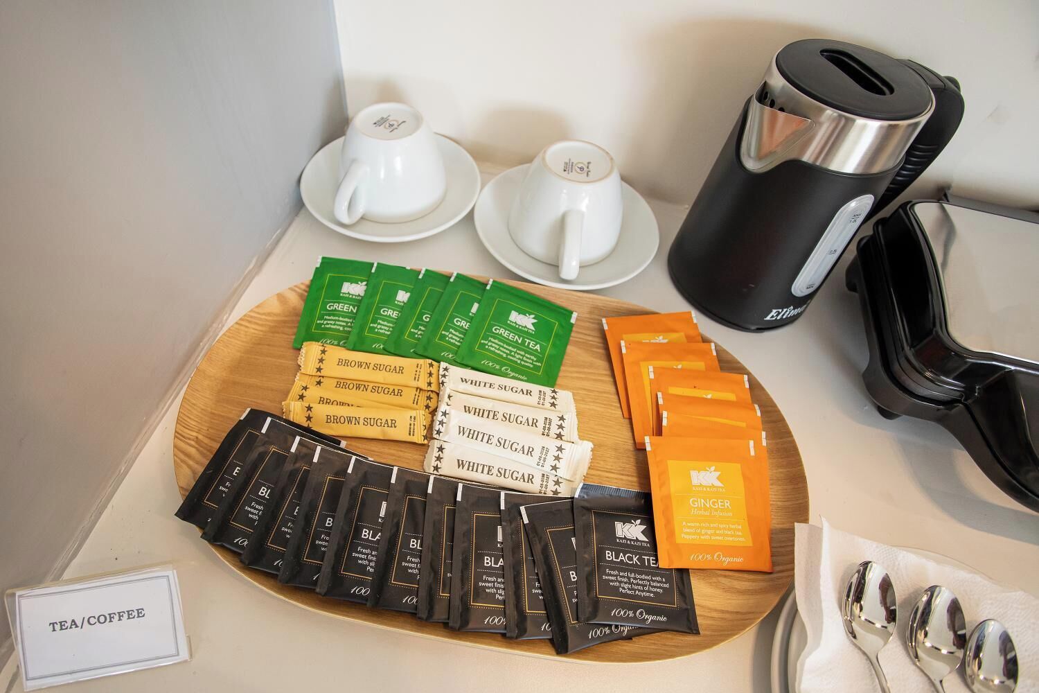 A tea/coffee station with various tea bags, two white cups, and a black coffee maker.