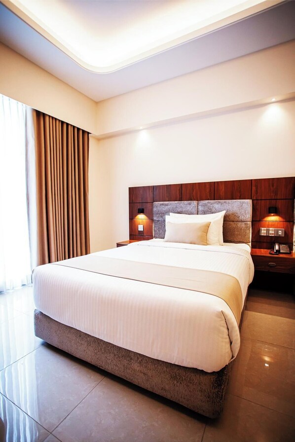 A hotel room with a large bed, wooden headboard, two bedside tables with lamps, and a large window with curtains. - The Royal Palace Uttara (Dhaka)