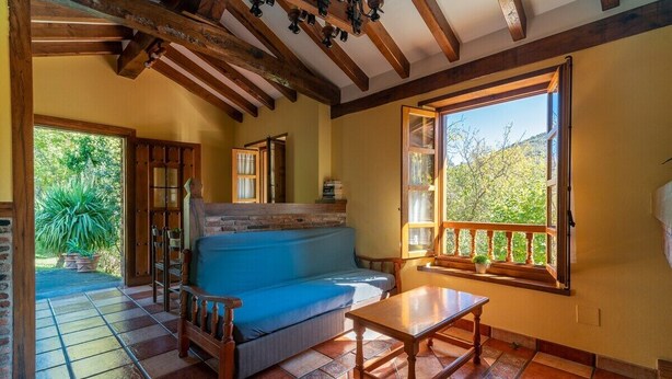 Ein gemütliches Zimmer mit Holzbalkendecke, einem blauen Sofa, einem Holztisch und einem Fenster mit Blick auf Grünflächen.