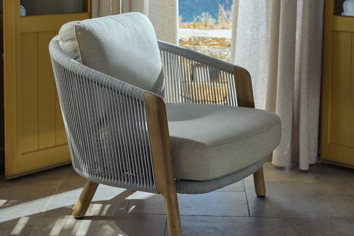 A chair with a woven backrest and wooden legs, positioned in a room with a view of the sea and mountains through large windows.