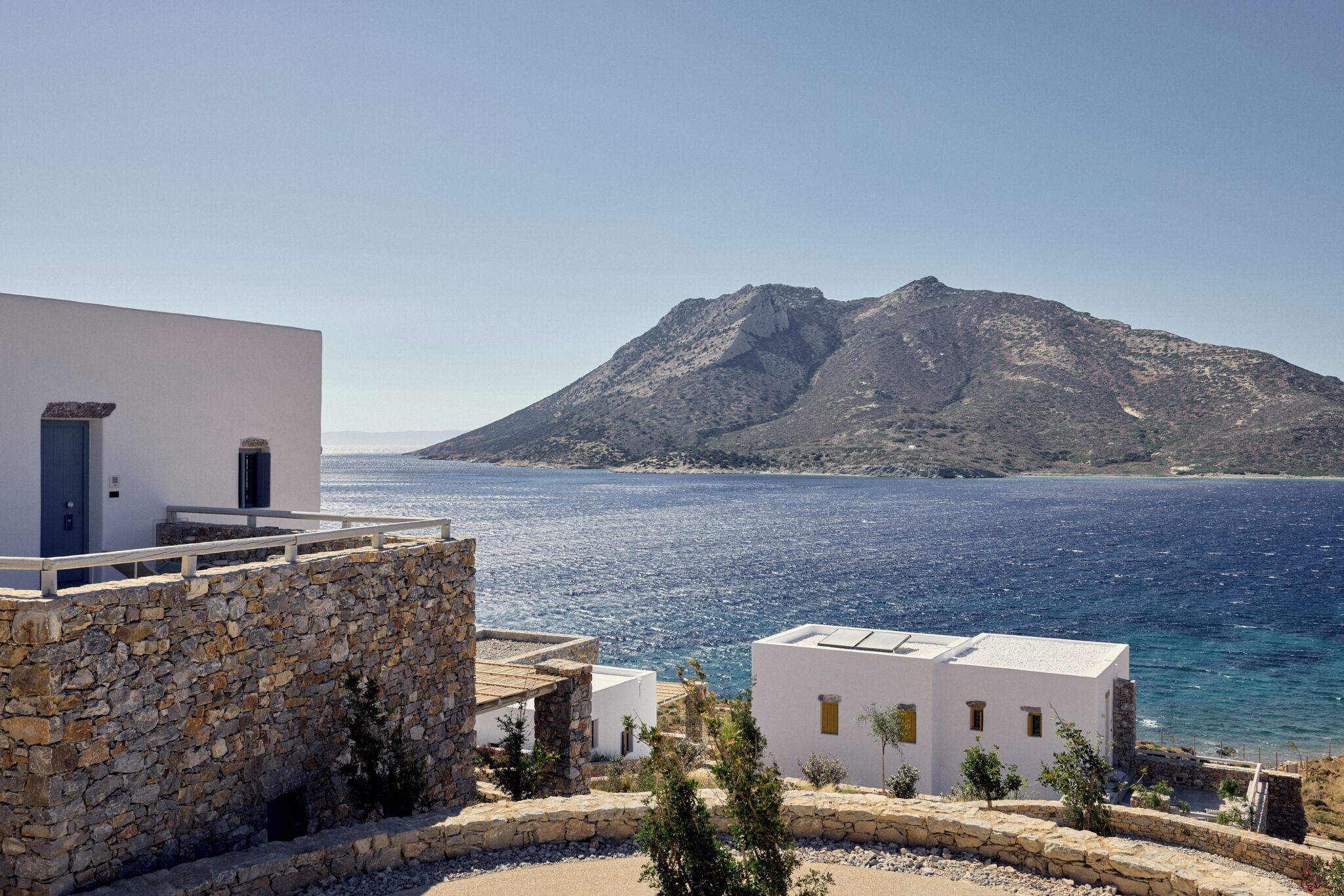 Une propriété située sur la côte, dotée d’un mur en pierre, de bâtiments blancs et offrant une vue sur la mer et une montagne.