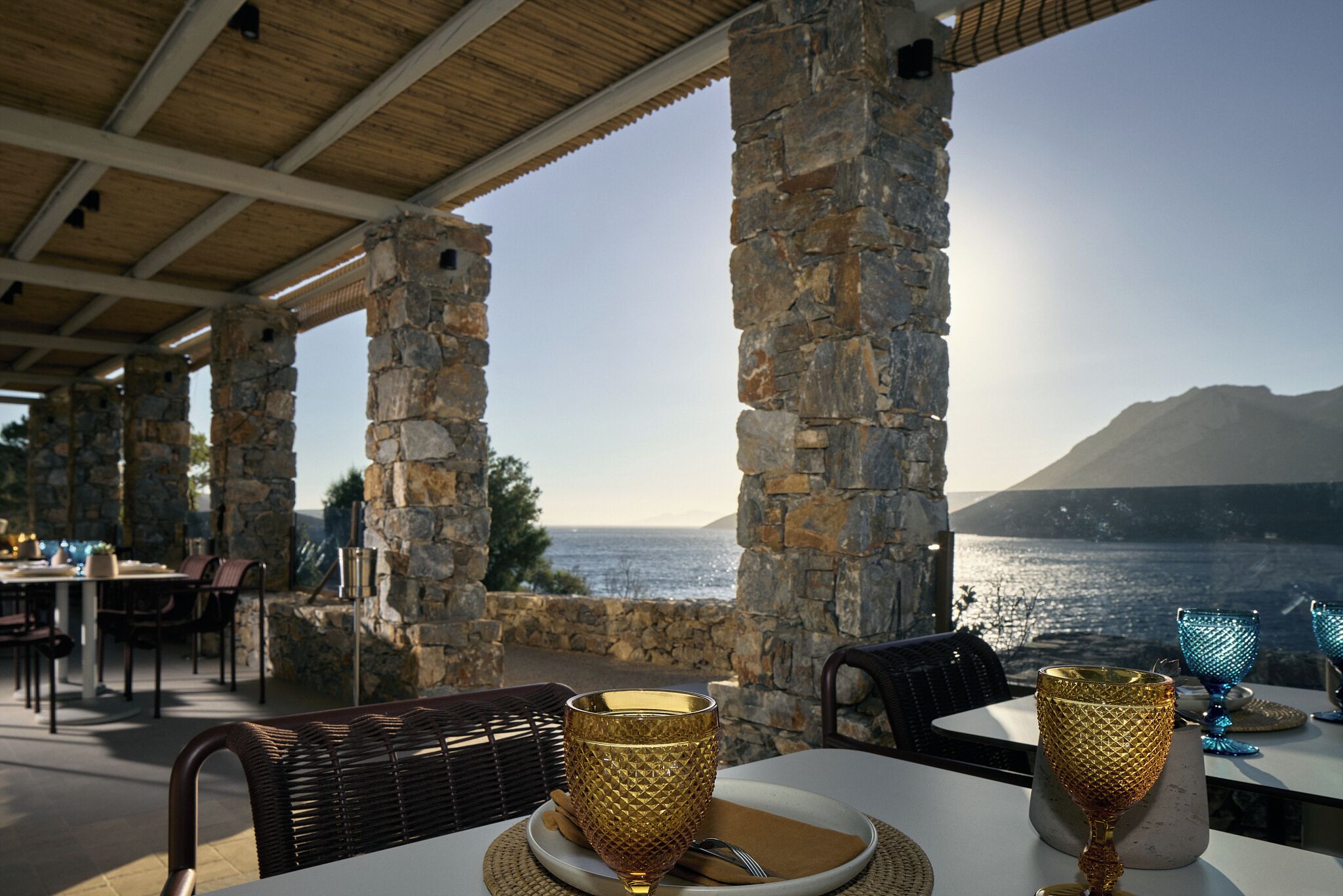 An outdoor dining area with stone pillars, tables, and chairs, overlooking a body of water and a mountain.