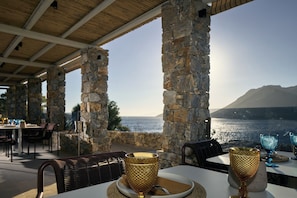 An outdoor dining area with stone pillars, tables, and chairs, overlooking a body of water and a mountain. - Amorgina (Amorgos)