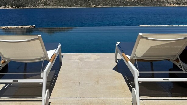 A poolside area with two lounge chairs and umbrellas, overlooking a body of water and a mountainous landscape. - Amorgina (Amorgos)
