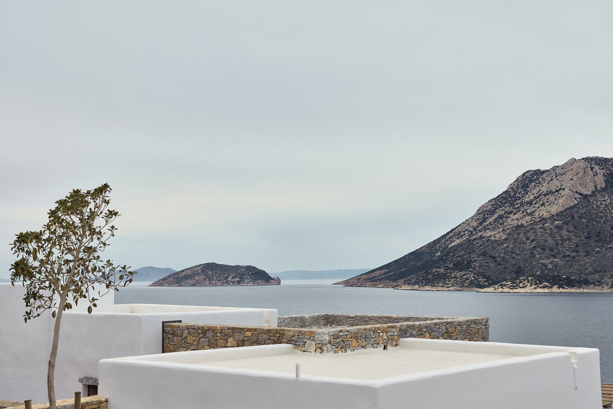 Una terraza en la azotea con vista al mar y a las montañas, paredes blancas y un árbol.