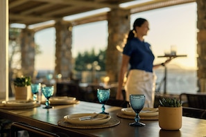 A restaurant setting with tables set for dining, a waiter in the background, and a view of the outdoors. - Amorgina (Amorgos)