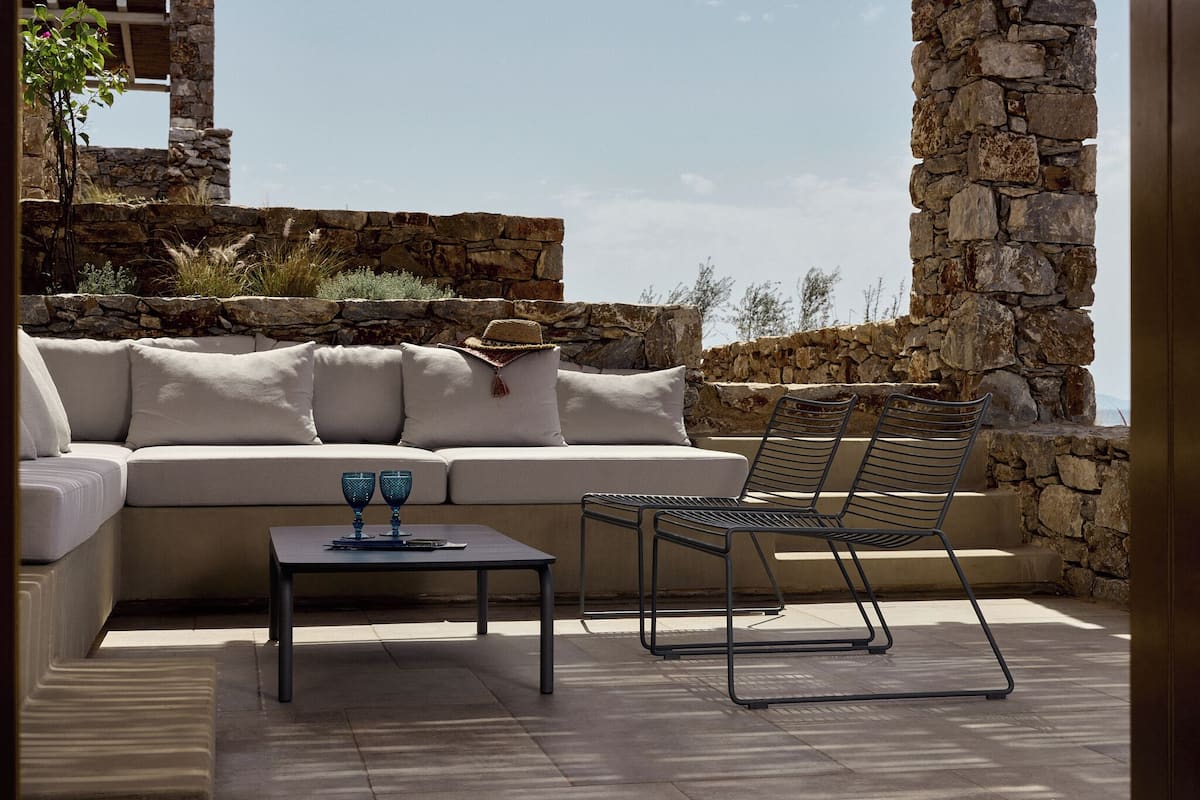 A sunlit outdoor seating area with a sofa, a metal table, and a metal chair, set against a stone wall and a clear sky.