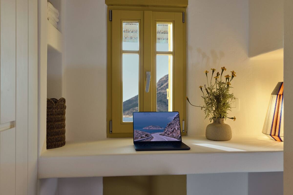 A laptop on a shelf with a window view, a vase with flowers, and a lamp.