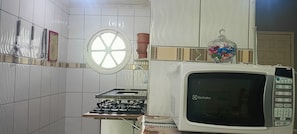 A kitchen with a gas stove, a microwave, and a window with a decorative wheel. - Cozy Grandma's Little House - 200m from the Sea + Pool + Pet-Friendly (Itanhaém)