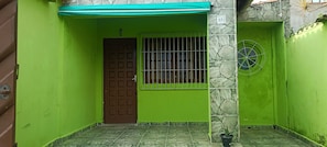 Un edifici amb parets verdes, una porta marró, una finestra amb reixes i una planta en test.