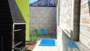 Een smalle buitenruimte met een barbecue van baksteen, een tafeltje met een plant en kleurrijke stoelen tegen een betegelde muur.