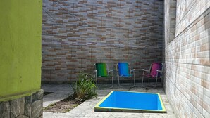 Een klein buitengebied met een blauw zwembad, een bakstenen muur en drie kleurrijke stoelen.