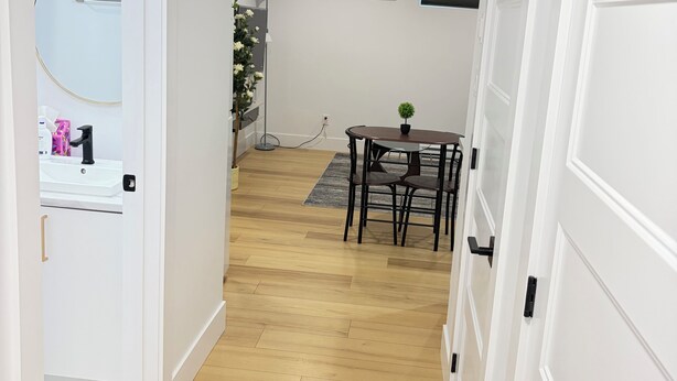 A hallway with a wooden floor, a white door, a small table with chairs, a potted plant, and an air conditioning unit on the wall. - Luxury 1 BedRoom Suite | New | Quite | Private| Near Amenities.  (Moncton)
