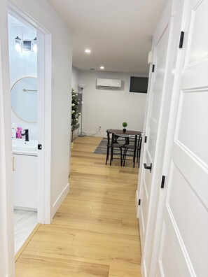 Een hal met een houten vloer, een witte deur, een kleine tafel met stoelen, een bloempot en een airconditioner aan de muur.