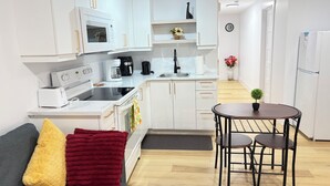 A compact kitchen with white cabinets, a dining table with chairs, a small potted plant, and a sofa with a yellow and red blanket. - Luxury 1 BedRoom Suite | New | Quite | Private| Near Amenities.  (Moncton)