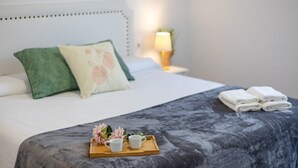 Una cama bien hecha con una manta gris, almohadas, una bandeja con tazas y flores, y toallas dobladas a un lado.