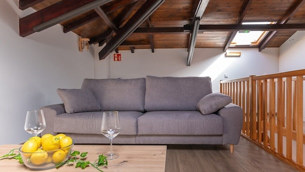 A modern living room with a grey sofa, wooden beams, a wooden railing, and a table with lemons and glasses. - Infinity Coast VI by Fidalsa (Miengo)