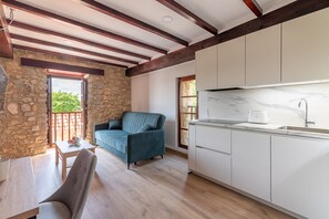 A modern kitchen with white cabinets and a marble countertop, a stone wall, and a blue sofa in a bright room with wooden beams. - Infinity Coast VI by Fidalsa (Miengo)