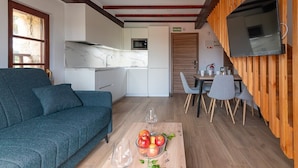 A modern living room with a sofa, a dining table with chairs, a kitchen area, and a television mounted on a wooden panel. - Infinity Coast VI by Fidalsa (Miengo)